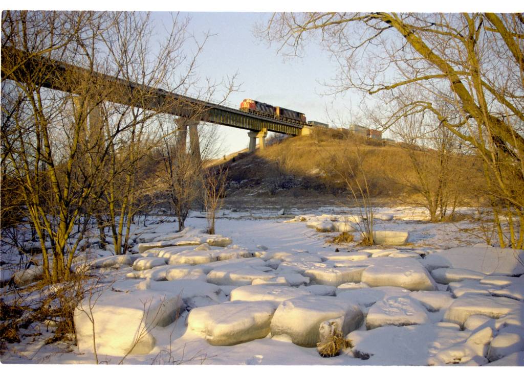 A CN Train begins crossing the trestle over the Humber River at Steeles and Islington area of North York, Ontario. Large ice pans are scattered outside the banks of the Humber River from when the river had earlier ice-jammed. The evening sun bathes the landscape in a golden light.